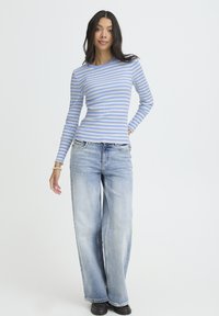 Gestreiftes Langarmoberteil in Hellblau und Beige, kombiniert mit ausgewaschenen blauen Jeans mit weitem Bein. Der Stoff wirkt gerippt; das Outfit wird vor einem schlichten Hintergrund präsentiert.