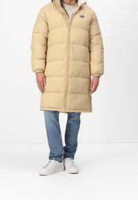 Beige lang pufferjakke med quiltet design, fuld lynlås og høj krave. Har elastiske manchetter. Kombination med blå jeans og hvide sneakers.