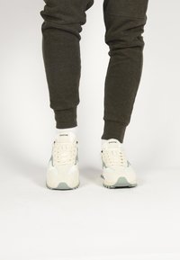 Witte en lichtgroene sneakers met een bovenwerk van suede en mesh, voorzien van veters en een gestructureerde zool, gedragen met donkergrijze joggers en witte sokken.