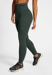Leggings taille haute de couleur vert foncé avec texture côtelée et logo au niveau de la taille, associés à des chaussures de sport noires et vertes.