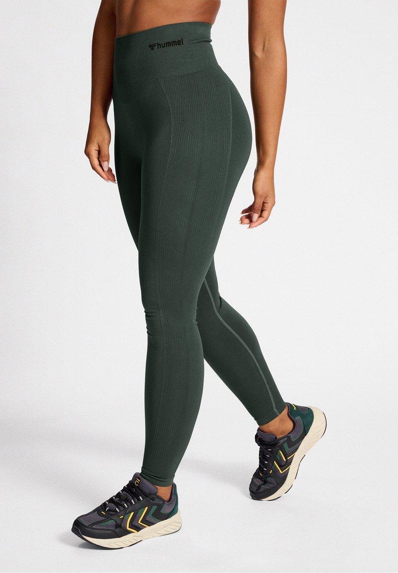 Leggings taille haute de couleur vert foncé avec texture côtelée et logo au niveau de la taille, associés à des chaussures de sport noires et vertes.