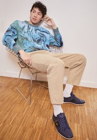 Sweter z dzianiny z niebiesko-zielonym marmurkowym wzorem, beżowe spodnie sztruksowe i granatowe trampki, zestawione z białymi skarpetkami, siedzące na metalowym krześle.