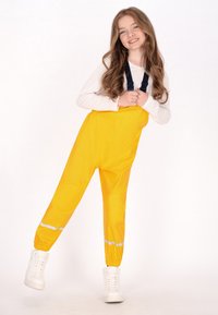 Usmievajúca sa mladá dievčina s dlhými vlasmi, oblečená v bielej dlhoprstej košeli, jasne žltých reflexných dažďových nohaviciach s čiernymi traky a bielych teniskách.