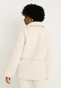 Chaqueta de forro polar color crema con una textura suave, cuello puntiagudo y un corte relajado. Presenta una costura en la cintura y mangas largas.