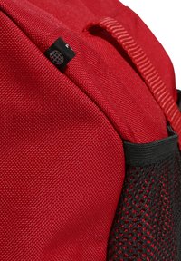 Mochila roja de tela con superficie texturizada, bolsillo lateral de malla negra y una pequeña etiqueta. Presenta un asa de transporte en la parte superior.
