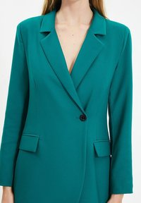 Blazer bleu sarcelle avec revers crantés, fermeture à un bouton noir, et deux poches avant. Tissu lisse avec coupe structurée et manches longues.