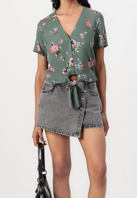 Camicetta floreale verde a maniche corte con vita annodabile, abbinata a una mini gonna in denim grigia con chiusura a bottoni frontale e orlo angolato.