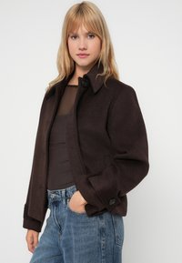 Chaqueta de mezcla de lana marrón con cuello, dos bolsillos frontales y cierre de botón. Combinada con una blusa ceñida negra y jeans azules.