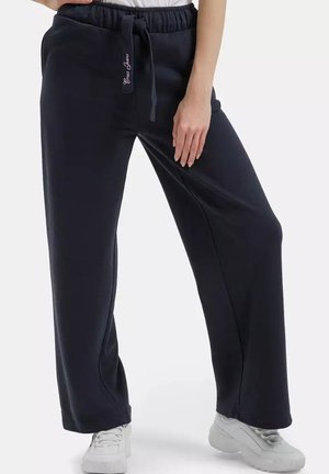 Osoba ubrana w czarne spodnie z szerokimi nogawkami i sznurkiem oraz białe trampki, stojąca z jedną ręką opartą na udzie.