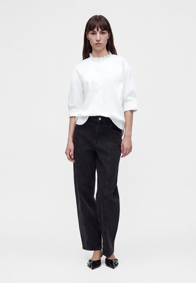 Femme portant un chemisier blanc à manches trois-quarts et un jean noir taille haute, debout sur un fond blanc.