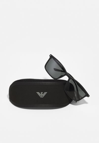 Ochelari de soare negri cu rame pătrate, având lentile întunecate, așezați pe un etui negru cu fermoar, cu un logo subtil în argintiu.