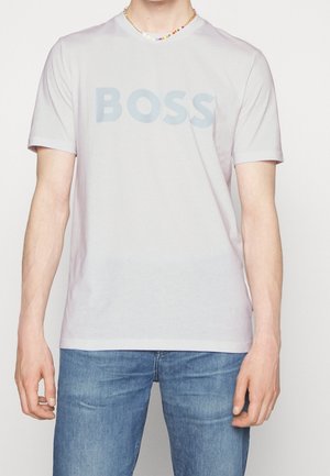 Persoon die een wit T-shirt met korte mouwen draagt met "BOSS" in grote letters op de borst gedrukt en een blauwe spijkerbroek.