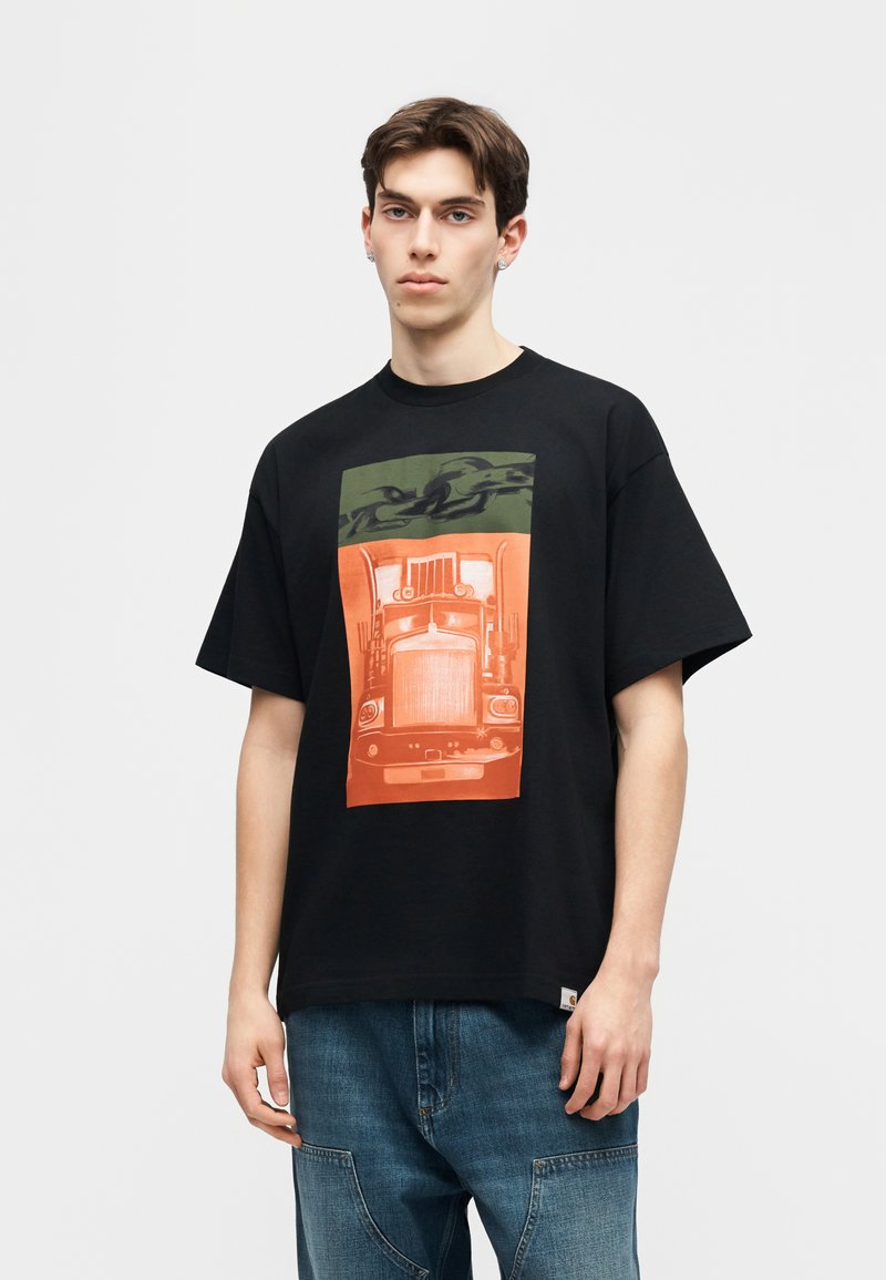 Giovane uomo che indossa una maglietta nera oversize con una grafica verde e arancione della parte anteriore di un camion e jeans blu, in piedi davanti a uno sfondo neutro.