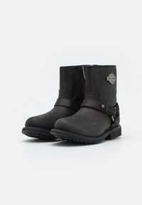 Botas de cuero negras hasta el tobillo con cierre de cremallera, detalle de correa y acento metálico con marca. Suela robusta con banda de rodadura texturizada para mayor tracción.