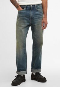 Jeans de denim em gradiente de azul claro a escuro, com um corte descontraído, costuras visíveis, bainhas dobradas e bolsos frontais. Usados com sapatos pretos.