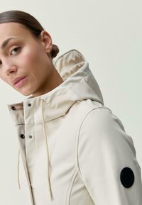 Béžová nepremokavá bunda s vysokým golierom, kapucňou na šnúrku a čiernou nášivkou loga na rukáve; hladká textura s minimálnym šitím.