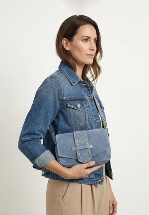 Kobieta w dżinsowej kurtce trzyma jasnoniebieską zamszową torebkę na ramię ze złotymi ćwiekami, stojąc na jednolitym jasnym tle.