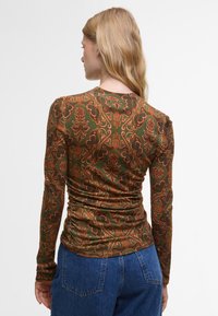 Langermige groene top met ingewikkeld oranje bloemenpatroon, textuurstof en geplooide details aan de achterkant, gecombineerd met blauwe denim jeans.