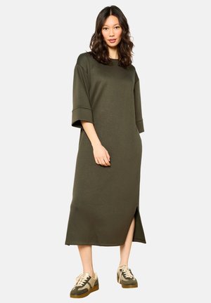 Olijfgroene, loszittende midi-jurk met korte mouwen en zijsplitten. Gemaakt van zacht materiaal, gecombineerd met tan kleurige en groene sneakers.