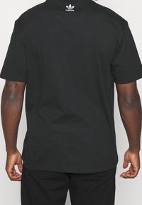 Svart bomullströja med rundad hals, korta ärmar och en liten vit Adidas-logotyp på bakhalsen. Slät textur.