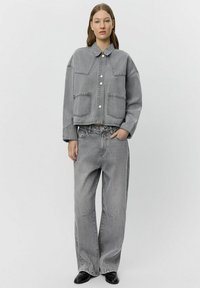 Giacca di jeans grigia tagliata, con ampie tasche frontali e bottoni a pressione, abbinata a pantaloni in jeans grigi larghi, caratterizzati da una sottile texture.