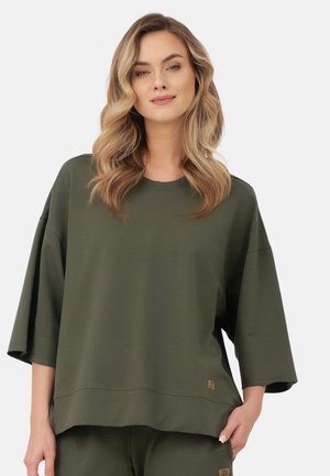 Kobieta o falowanych blond włosach, ubrana w oversize ciemnozielony krótki rękaw i pasujące spodnie na jednolitym tle.