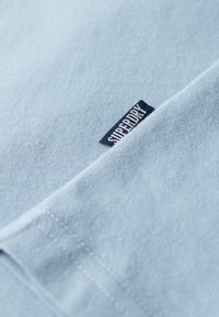 Lichtblauw katoen t-shirt met een gestructureerde oppervlakte en een klein donkerblauw label met de tekst "Superdry" aan de linkerkant, nabij de zoom.