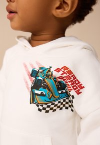 Balta bliuzonas su spalvinga animacinių lenktyninių automobilių grafika, su languotu raštu ir ryškiu tekstu, pagamintas iš minkštos medžiagos.