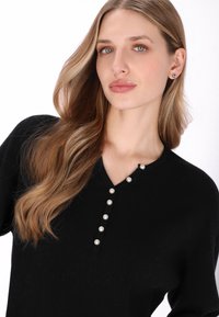 Black ribbed long-sleeve sweater featuring a V-neckline with a line of decorative pearl buttons for accent. Smooth texture and fitted design.