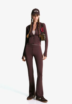 Femme avec des cheveux tressés portant une veste zippée marron foncé, un pantalon évasé assorti, des baskets, une casquette noire et portant un petit sac à main avec un clip rouge.
