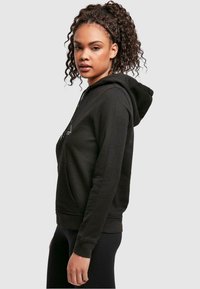 Black pullover hoodie featuring a front pocket, drawstring hood, and ribbed cuffs. Soft fabric with a relaxed fit, side view shown.