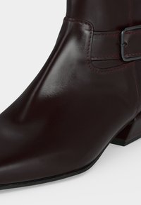 Bottine en cuir marron foncé avec bout pointu, petit talon carré et sangle avec boucle métallique autour de la cheville.