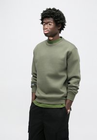 Φούτερ σε ελιά πράσινο με στρογγυλή λαιμόκοψη, ριμπ μανσέτες και ραφές στους ώμους. Φορεμένο πάνω από ένα σκούρο πράσινο πουκάμισο με στρώσεις. Μαύροι cargo παντελόνια.