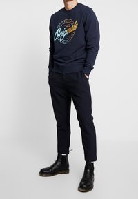 Marinblå sweatshirt med trycket "Originals"; ribbade muddar och fåll. Matchas med mörka byxor och svarta Chelsea-stövlar.