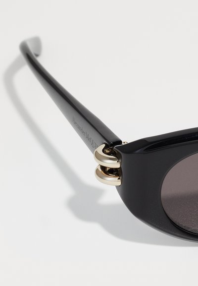 Occhiali da sole neri con finitura lucida, dotati di dettagli in metallo color oro e una distintiva finitura ad anello sulla tempia. Lenti scure.