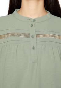 Blusa verde chiaro con scollatura rotonda, dotata di una fila di bottoni, rifiniture in pizzo decorative e dettagli plissè in vita.