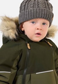 Grüne wasserdichte Jacke mit fellgefütterter Kapuze, grauer Strickmütze und Vorderreißverschlüssen. Sie verfügt über einen horizontalen weißen Streifen und Reißverschlüsse mit Holzakzenten.