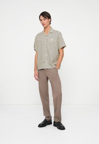 Kortebloemig, met kraag, shirt in beige met zwarte cirkelpatronen, gecombineerd met lichtbruine broek en zwarte schoenen.