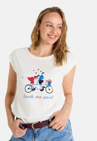 Camiseta blanca con un estampado gráfico de una bicicleta tándem azul que transporta a un hombre y una mujer, rodeados de corazones y flores. El texto debajo dice "Roule ma poule."