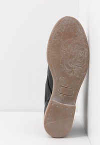 Chaussure en cuir noir avec une surface lisse et une semelle en caoutchouc brune texturée. Présente des détails gravés et une indication de taille 37.