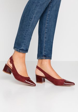Fødder i blå jeans iført burgunderfarvede slingback blokhæle med spidse, lakovertrukne tåspidser på en hvid gulv.