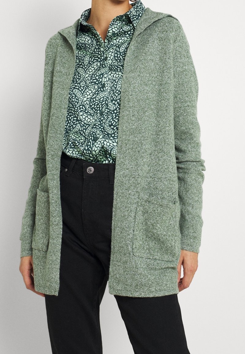 Světle zelený strukturovaný kardigan s kapucí, otevřený vpředu a s dvěma předními kapsami, nošený přes vzorovanou tmavě zelenou košili a černé kalhoty.