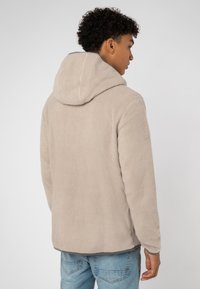 Beige Fleecejacke mit Kapuze, strukturierter Oberfläche und geschw emagtem Saum. Kombiniert mit hellblauen Jeans.