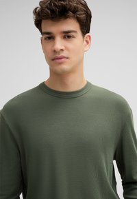 Langärmliges T-Shirt mit Rundhalsausschnitt aus strukturiertem olivgrünem Stoff. Bietet einen lockeren Schnitt und hat einen gerippten Halsausschnitt mit Nahtdetails.