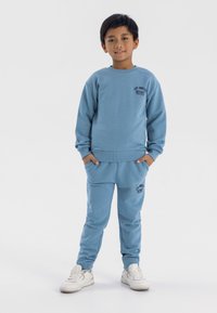 Blå bomullströja och matchande sweatpants med logotext på bröstet och låret. Har rund halsringning, ribbade ärmslut och fickor. Vita sneakers.