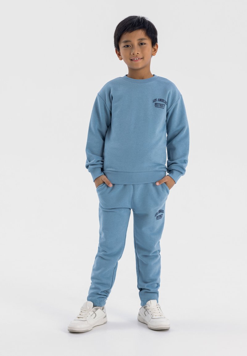 Blå bomullströja och matchande sweatpants med logotext på bröstet och låret. Har rund halsringning, ribbade ärmslut och fickor. Vita sneakers.