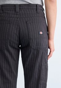 Pantaloni în dungi închise, realizați dintr-un material durabil, cu buzunare laterale, o etichetă cu logo pe spate și cusături vizibile.