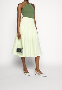 Débardeur côtelé vert olive associé à une jupe midi plissée jaune clair, des escarpins blancs à bout pointu, et un petit sac à main noir.