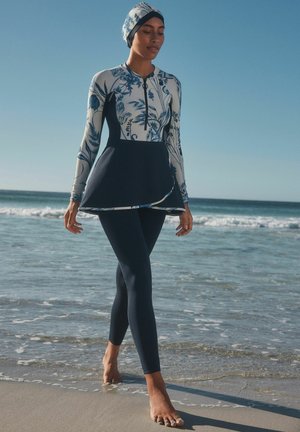 Ensemble de bain deux pièces comprenant un haut floral à manches longues avec fermeture éclair et un peplum bleu marine, associé à un legging ajusté bleu marine.