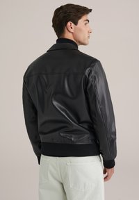 Chaqueta bomber de cuero negro con cuello y puños acanalados, con una textura suave y un ajuste entallado, llevada sobre pantalones de color claro.